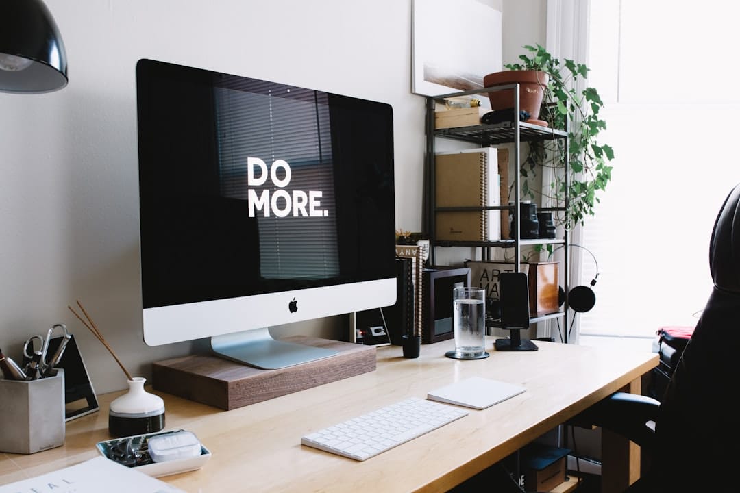 A minimalist and tidy workspace featuring an imac on a wooden stand with a motivational "do more." wallpaper, flanked by stationery and a potted plant, encouraging productivity and focus.