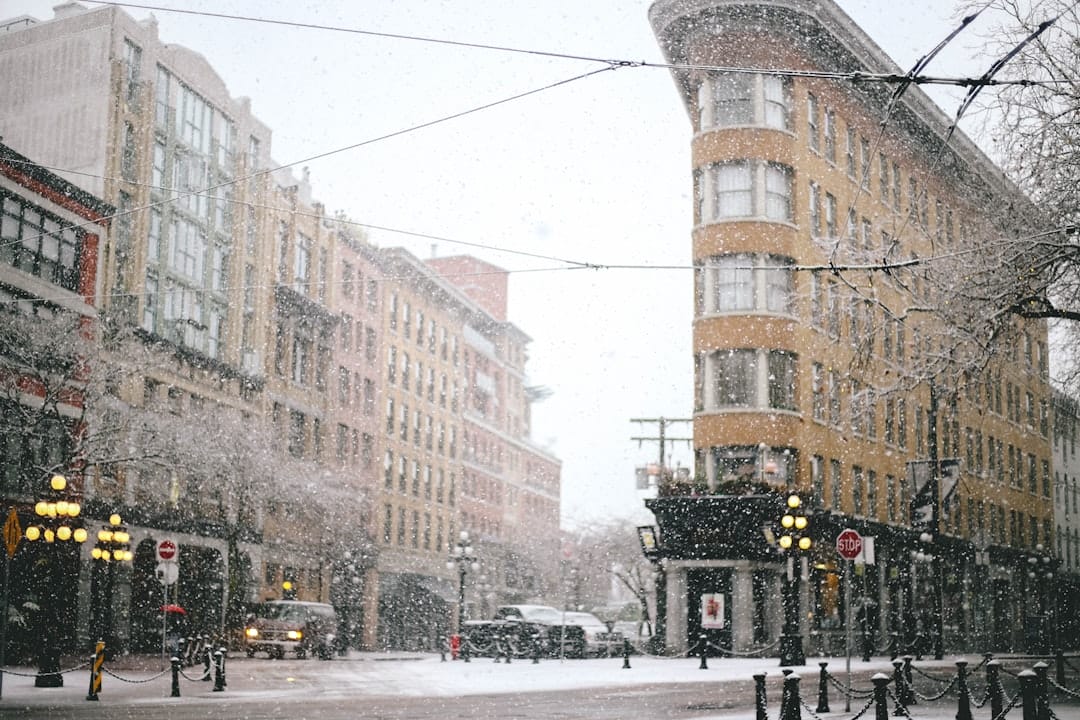 Snow gently falls on a city street, dusting historic buildings and street lamps with a soft, wintry blanket, while the world moves at a slower, quieter pace.