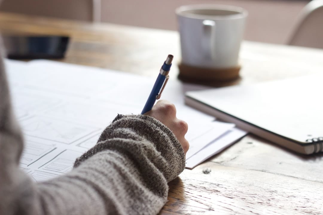 A Person In A Gray Sweater Is Writing Notes With A Blue Pen On Documents At A Wooden Table, With A Coffee Cup In The Background.