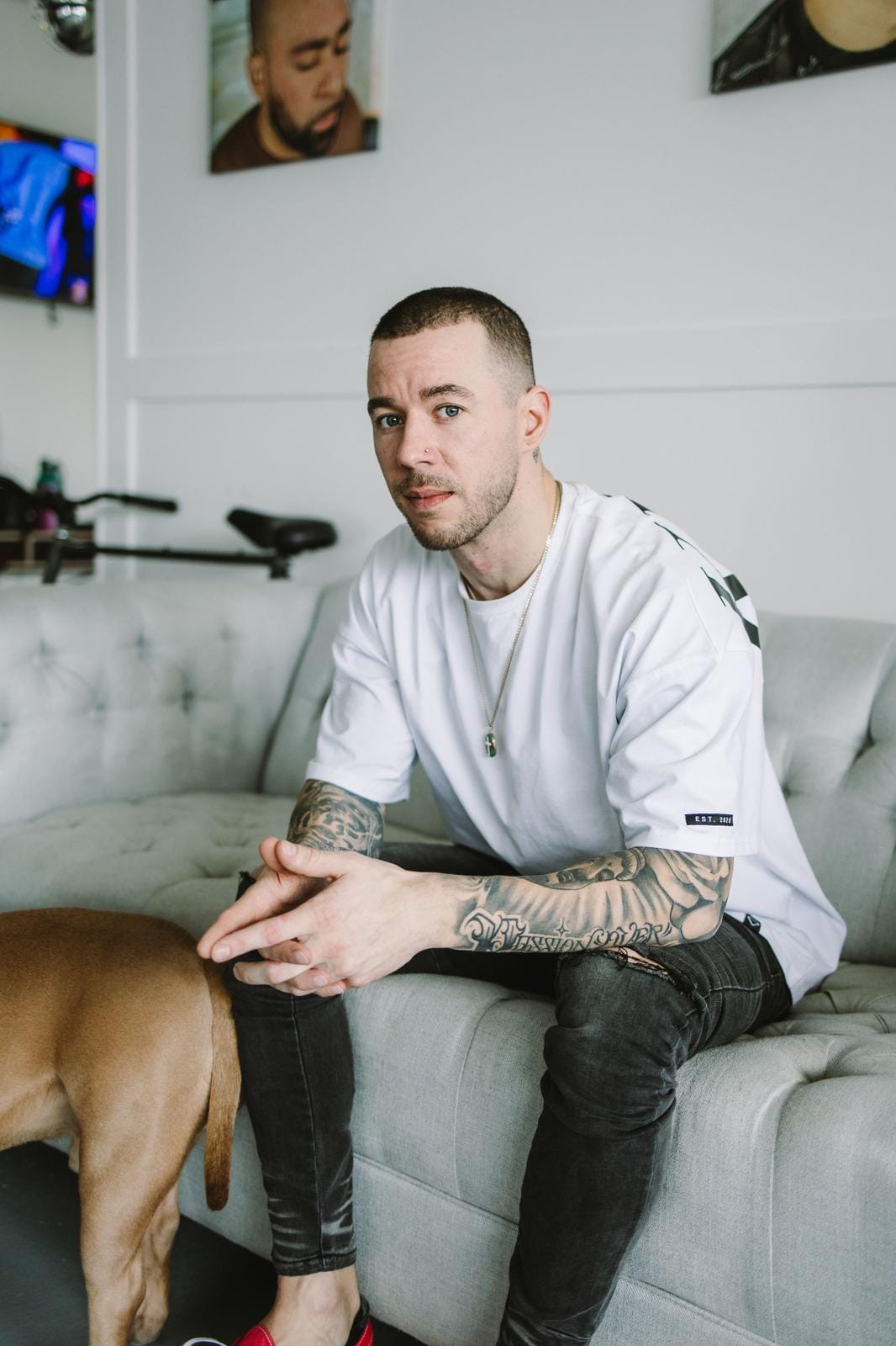 A Man With Tattoos On His Arms Sits On A Gray Couch, Looking Toward The Camera. He Is Wearing A White Shirt And Black Jeans. Part Of A Brown Dog's Body Is Visible Next To Him. The Background Shows Two Paintings And A Television.