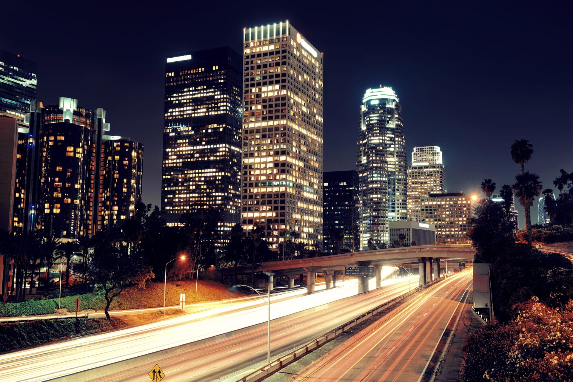 A Vibrant Nighttime Cityscape Featuring Illuminated Skyscrapers, A Glowing Freeway With Streaks Of Car Lights, And Dark, Clear Sky. The Urban Landscape Is Alive With Lights From High Rise Buildings And Cars, Creating A Dynamic And Bustling Atmosphere.